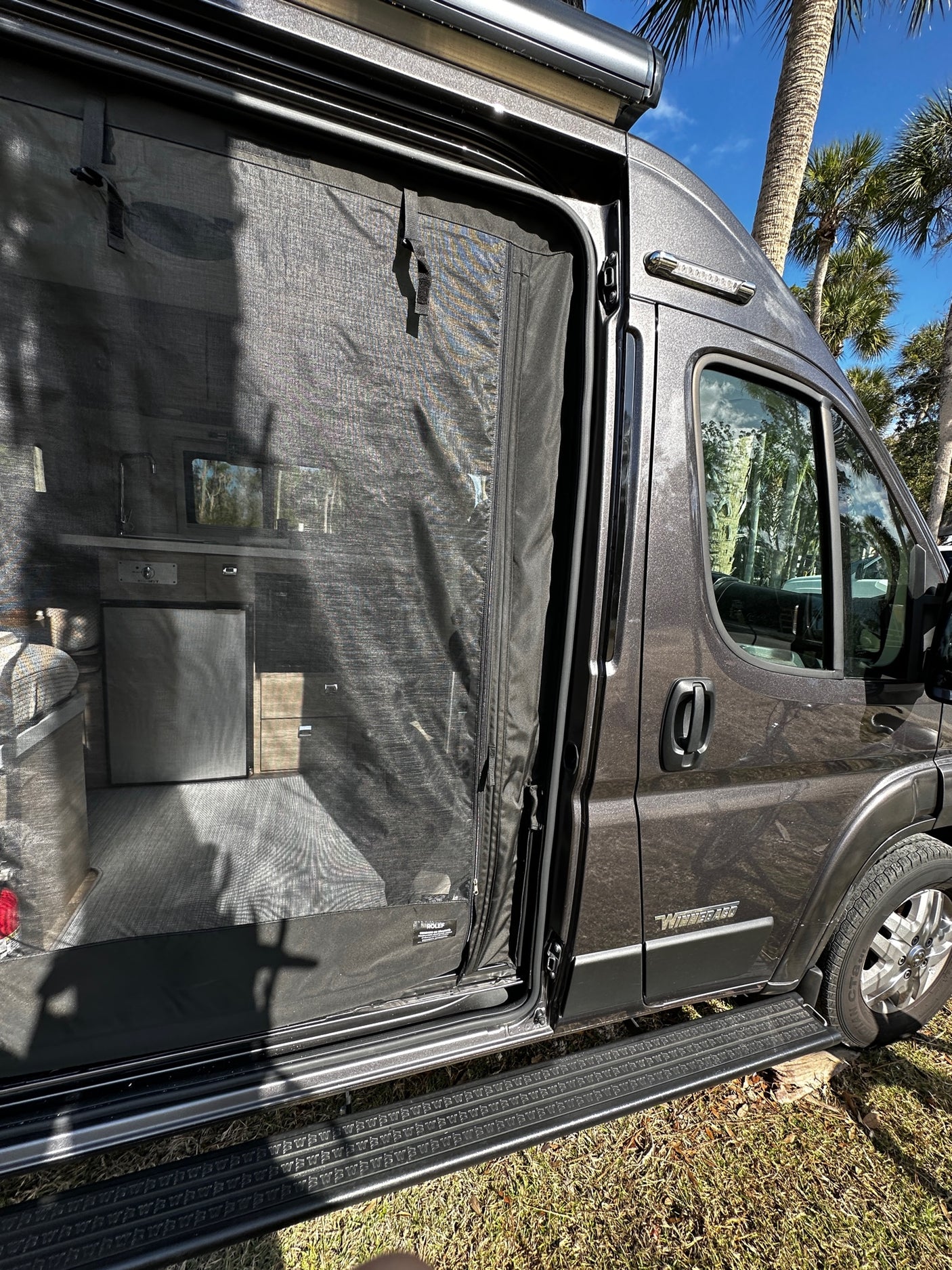 This grey Winnebago RV rental has its side door open, showcasing a mesh screen and interior details, with palm trees visible.