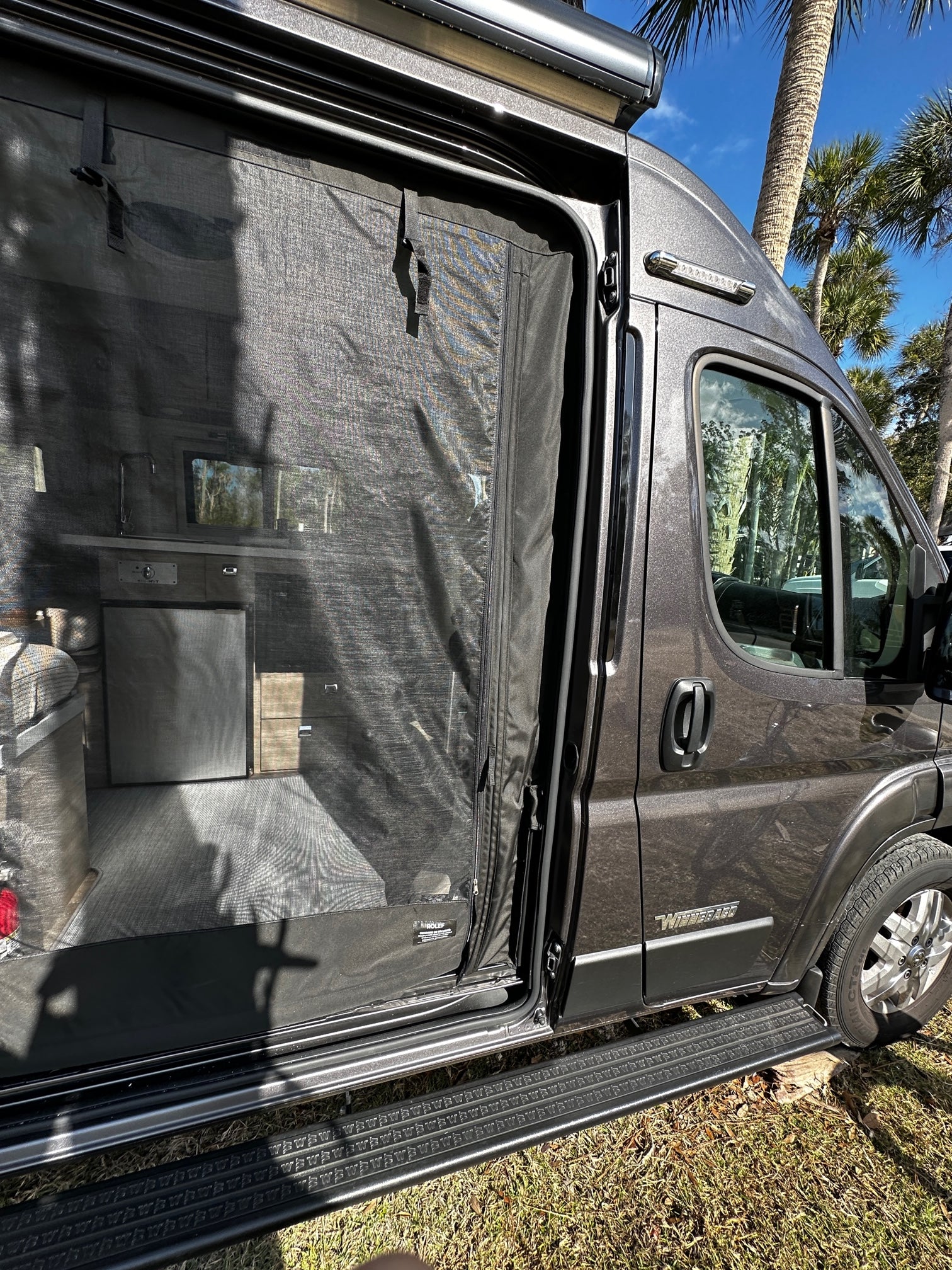 This grey Winnebago RV rental has its side door open, showcasing a mesh screen and interior details, with palm trees visible.