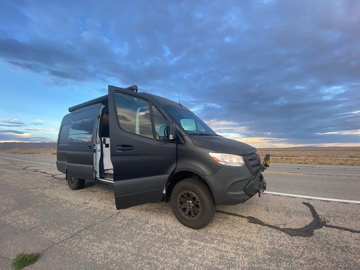 Adventure-ready matte black Sprinter van RV rental parked on a desert road with doors open under a cloudy sky.