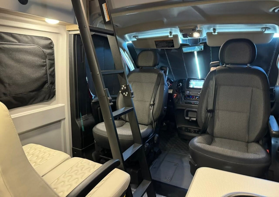 Inside an RV rental: view from the rear to the front cabin showing gray driver/passenger seats, dashboard, and an interior ladder.