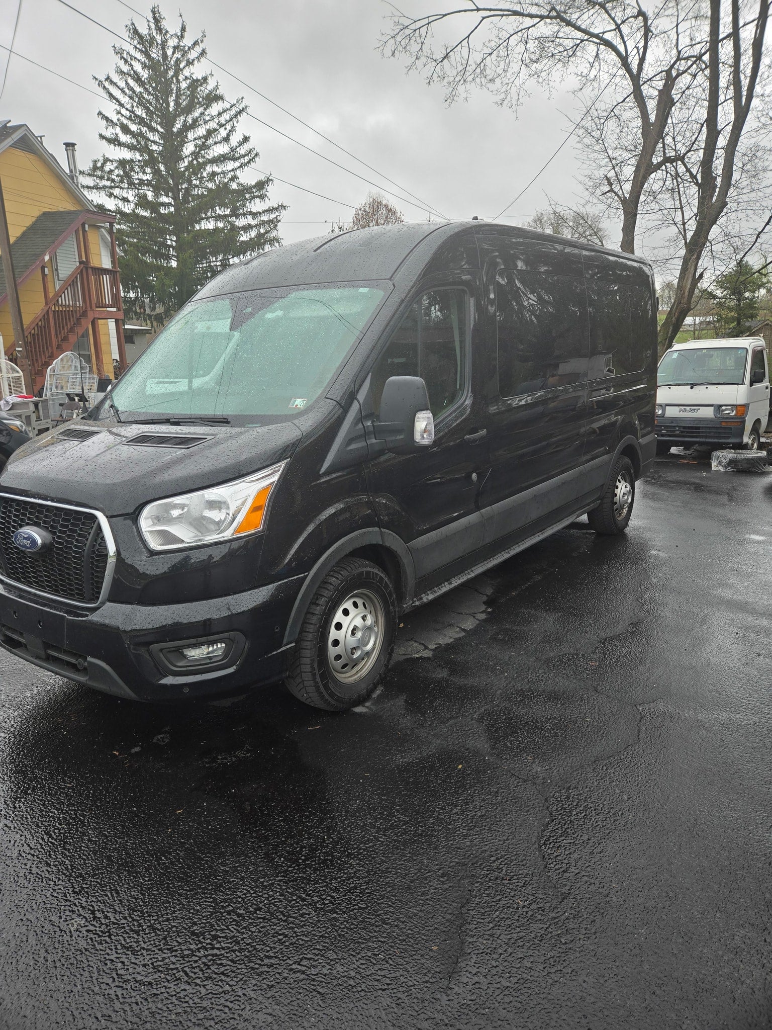 A black Ford Transit van, commonly used for RV rental conversions or commercial transport, sits on wet pavement. Another white van is behind.