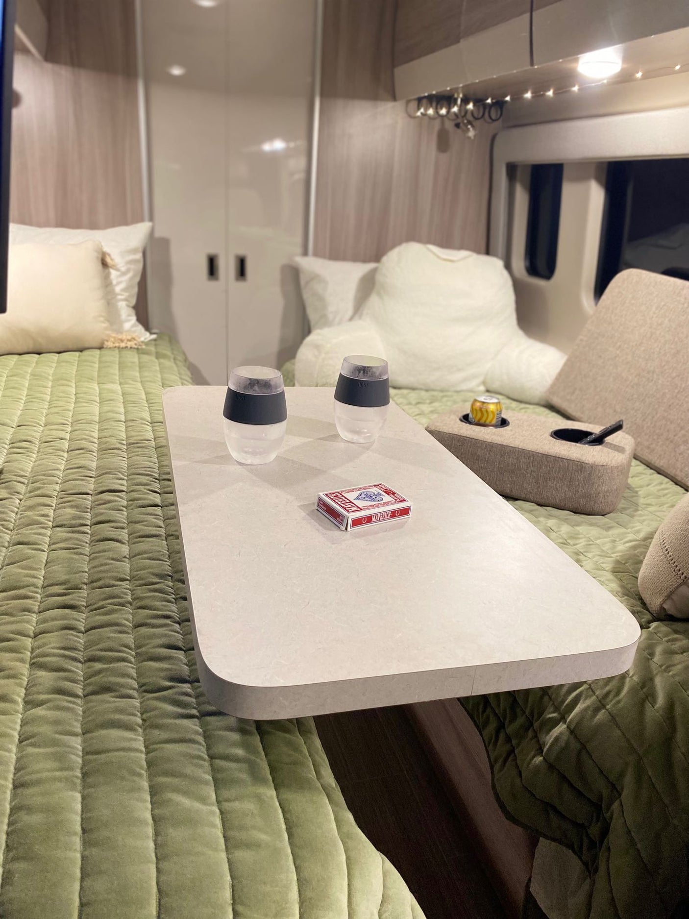 Inside a comfortable RV rental: two beds with plush green covers, a table for games/drinks, and overhead lights.