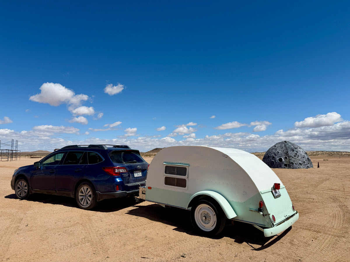 Blue car towing a light green teardrop rv rental in a vast desert landscape, featuring a large, cratered moon sculpture under a clear sky. — photo 1