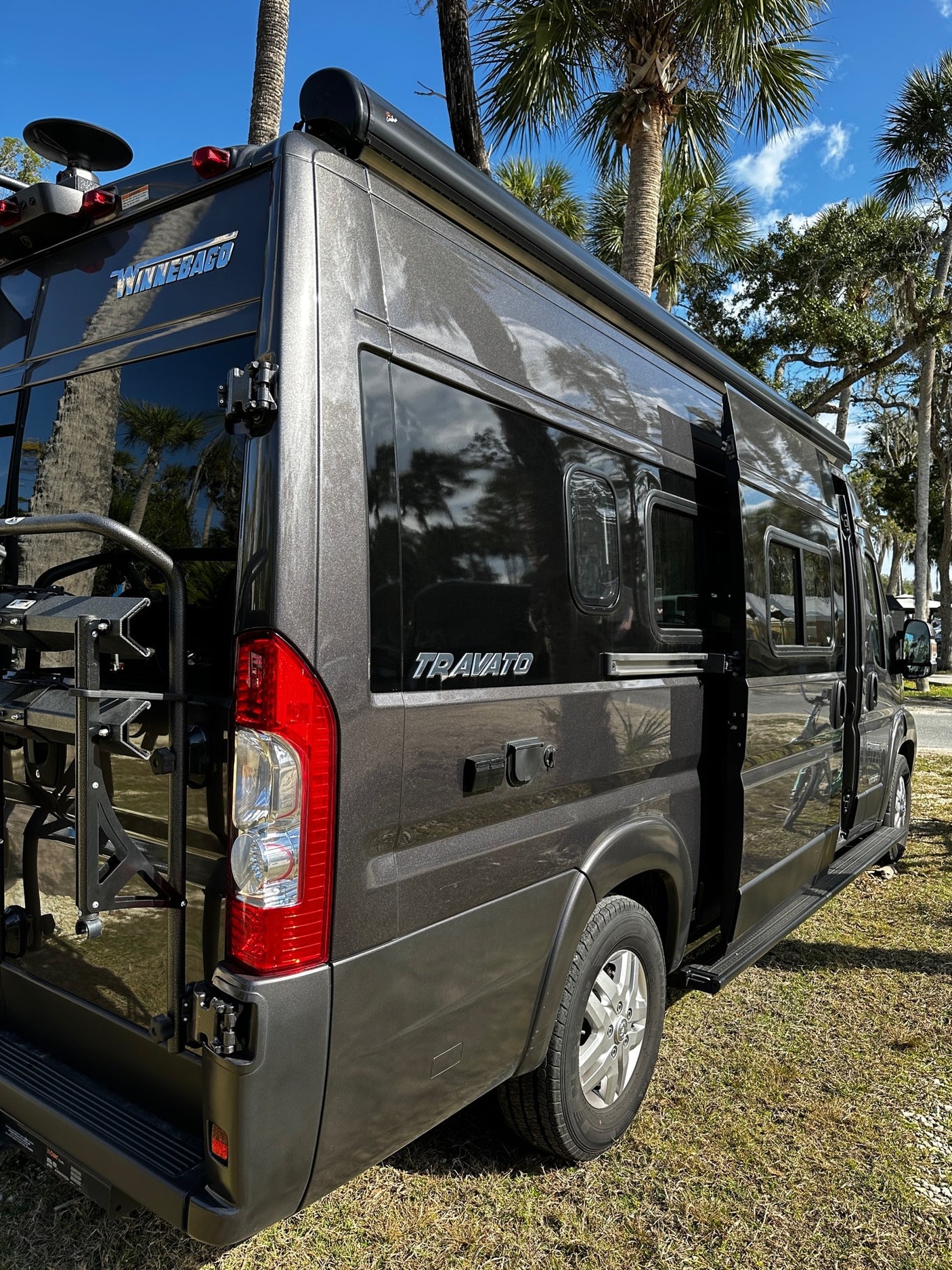 Dark grey Winnebago Travato rv rental. This camper van features an awning and rear rack, ideal for road trips.