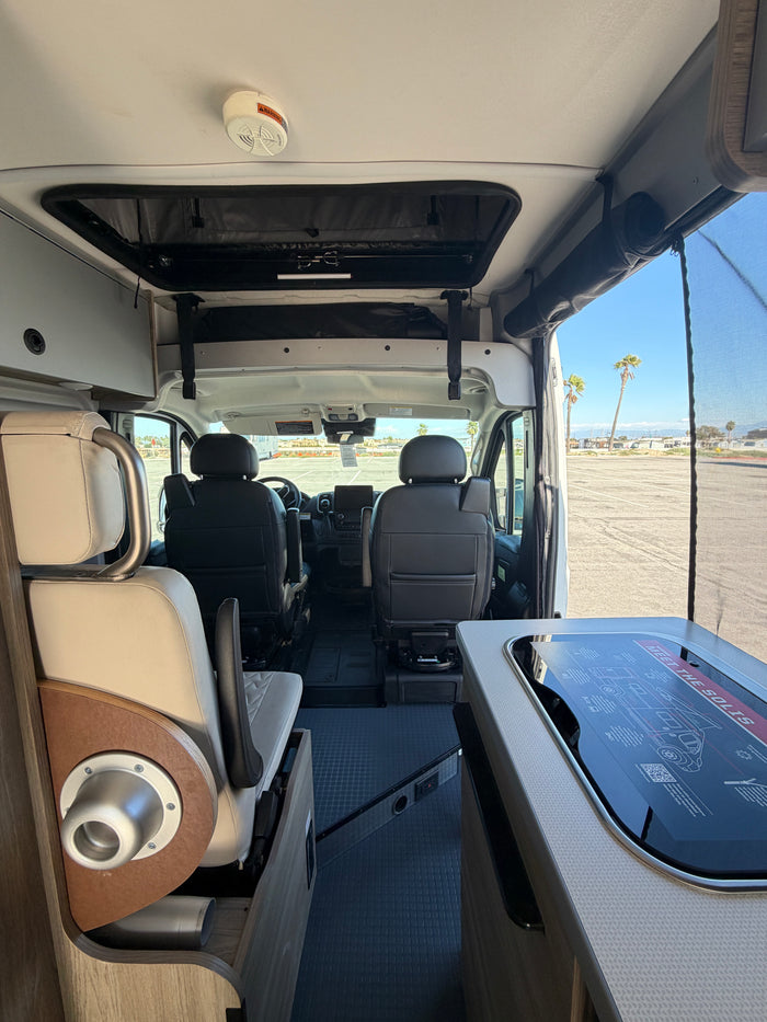 RV rental: Inside a Winnebago Solis camper van. View shows cockpit, kitchen, pop-top bed, with palm trees outside.