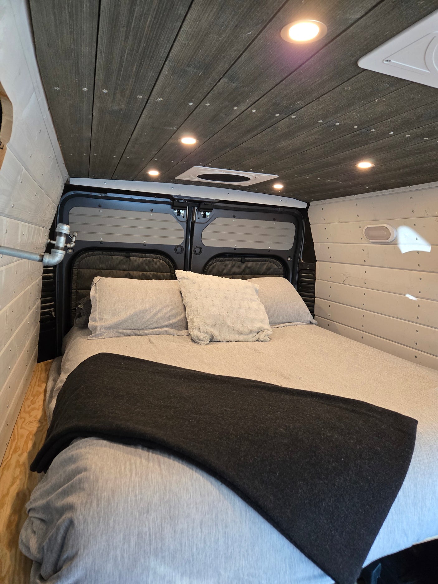 Cozy RV rental bed with grey bedding, dark wood ceiling with lights, and plank walls. Perfect for van life.