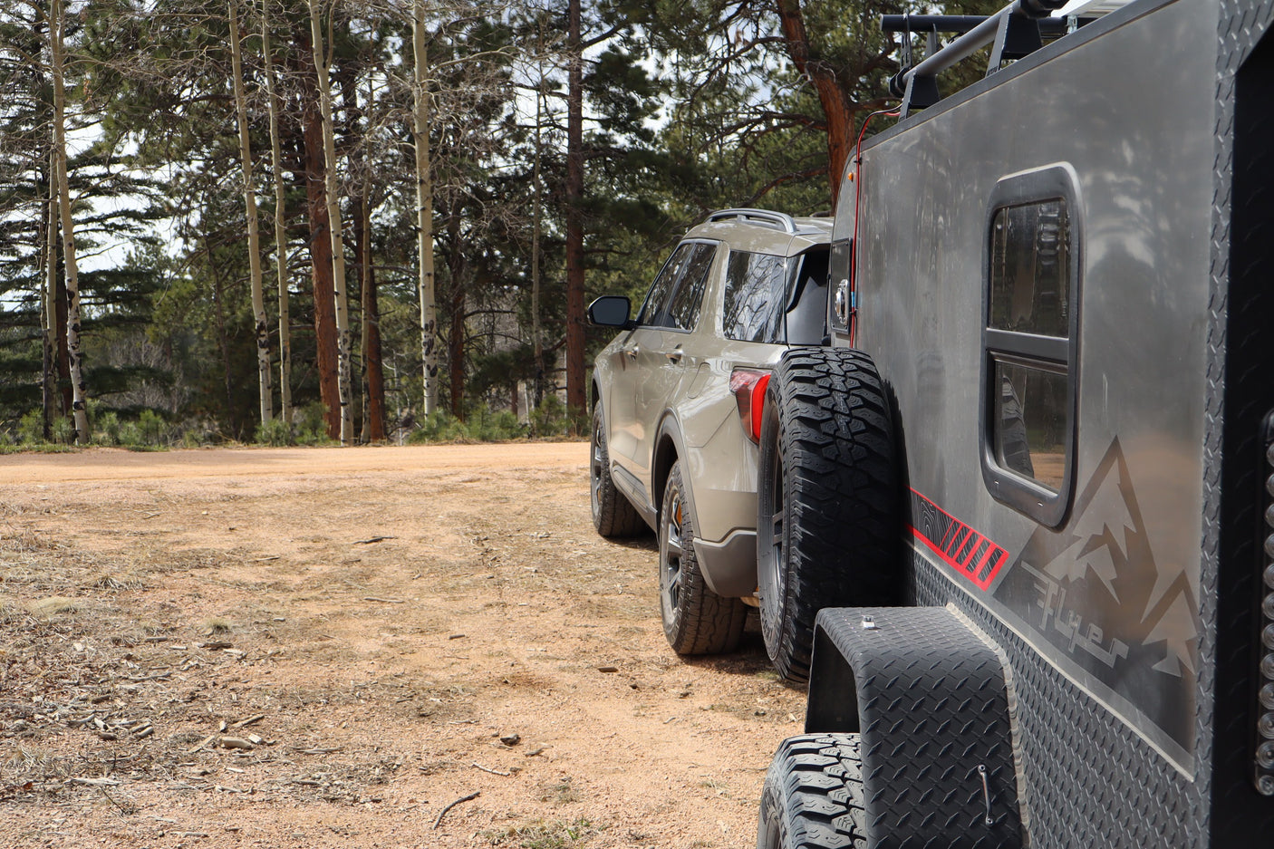 A tan SUV and gray overland trailer, ideal for an RV rental adventure, parked on a dirt road by pine and aspen trees.
