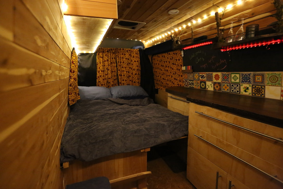 Cozy RV rental interior featuring a comfy bed, glowing wood paneling, and a colorful tiled kitchen with string lights.