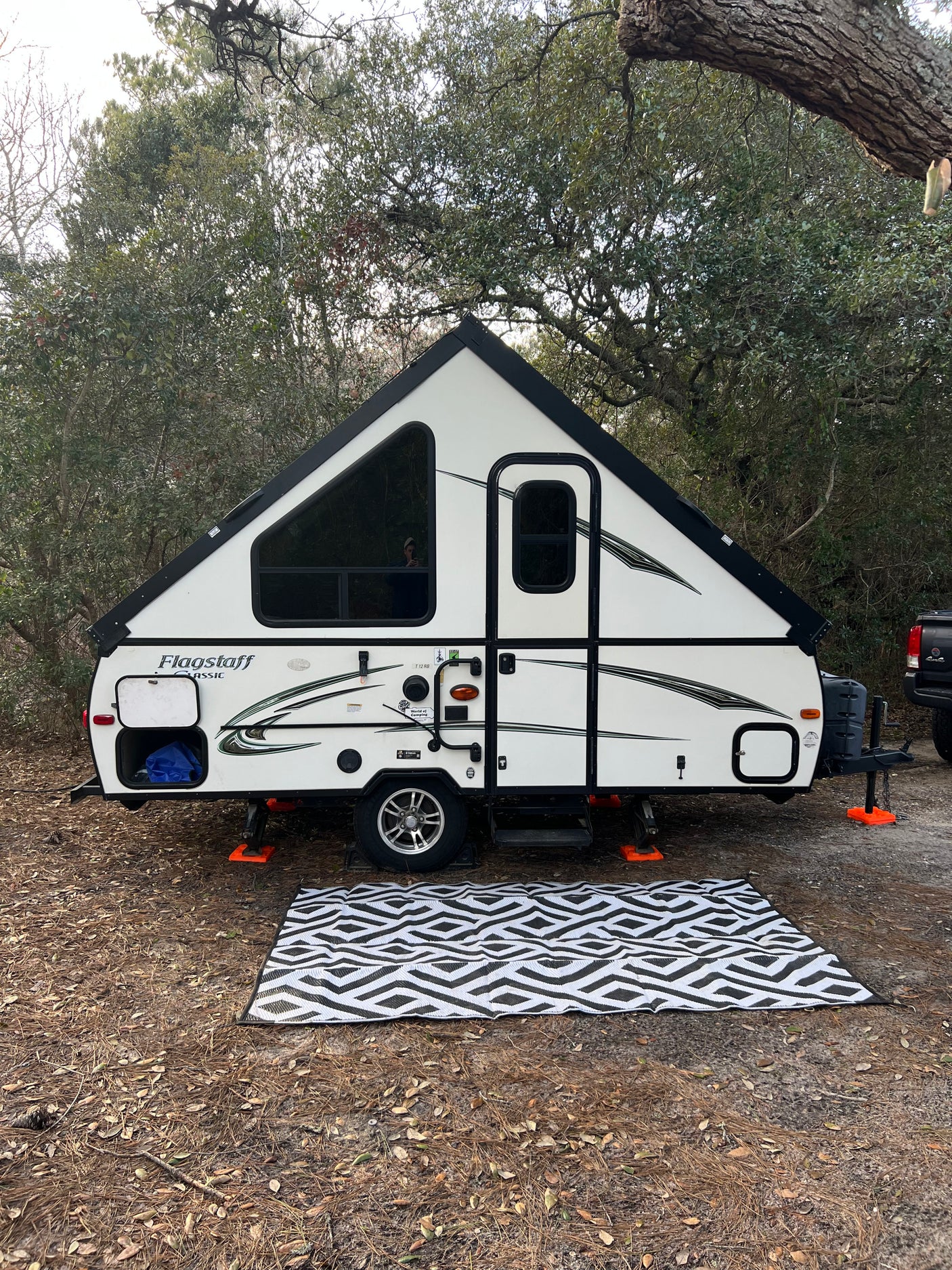 A white A-frame pop-up rv rental sits ready for camping on a pine needle forest floor with a patterned outdoor rug.
