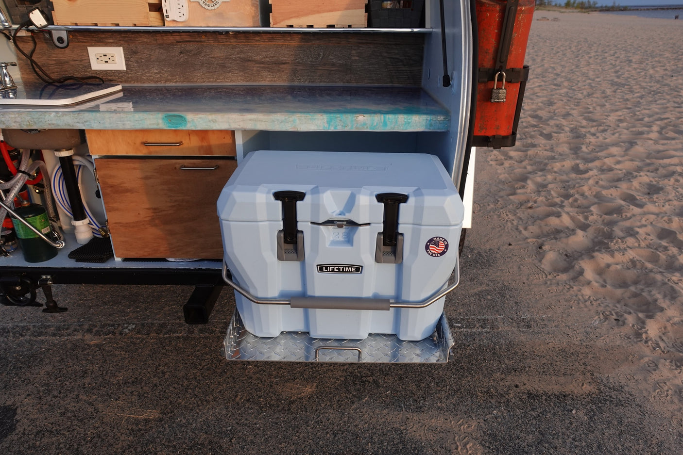 RV rental: Rear external view showing a light blue pull-out cooler, sink area, and storage. Parked by a sandy beach.