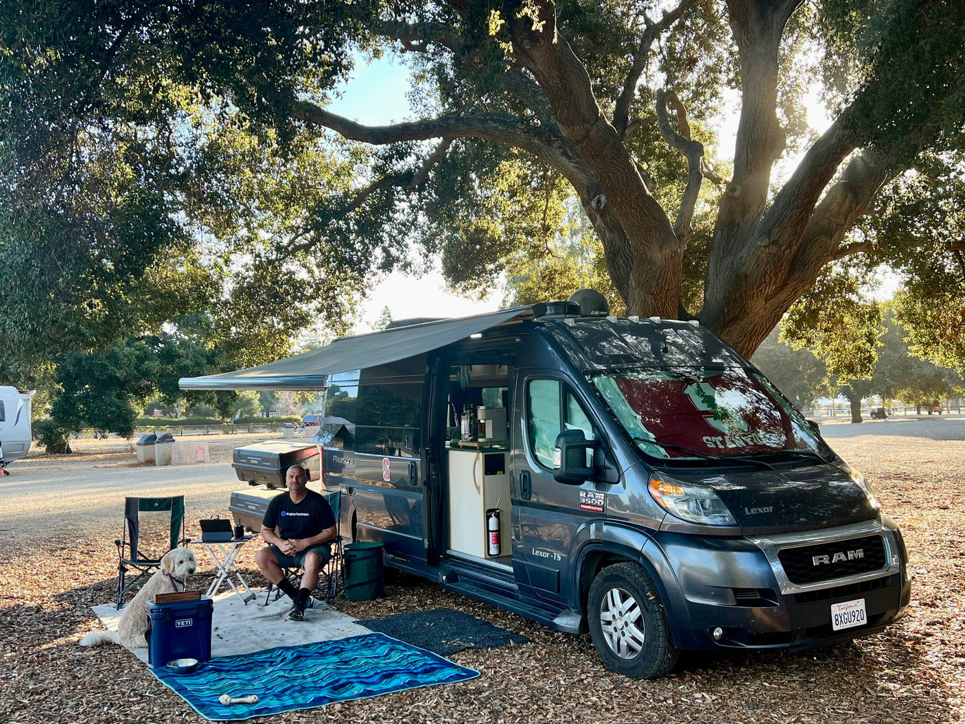 Man and dog relaxing outside their Pleasure-Way Lexor-TS RV rental (Ram ProMaster van) under a large shaded tree, enjoying outdoor camping.