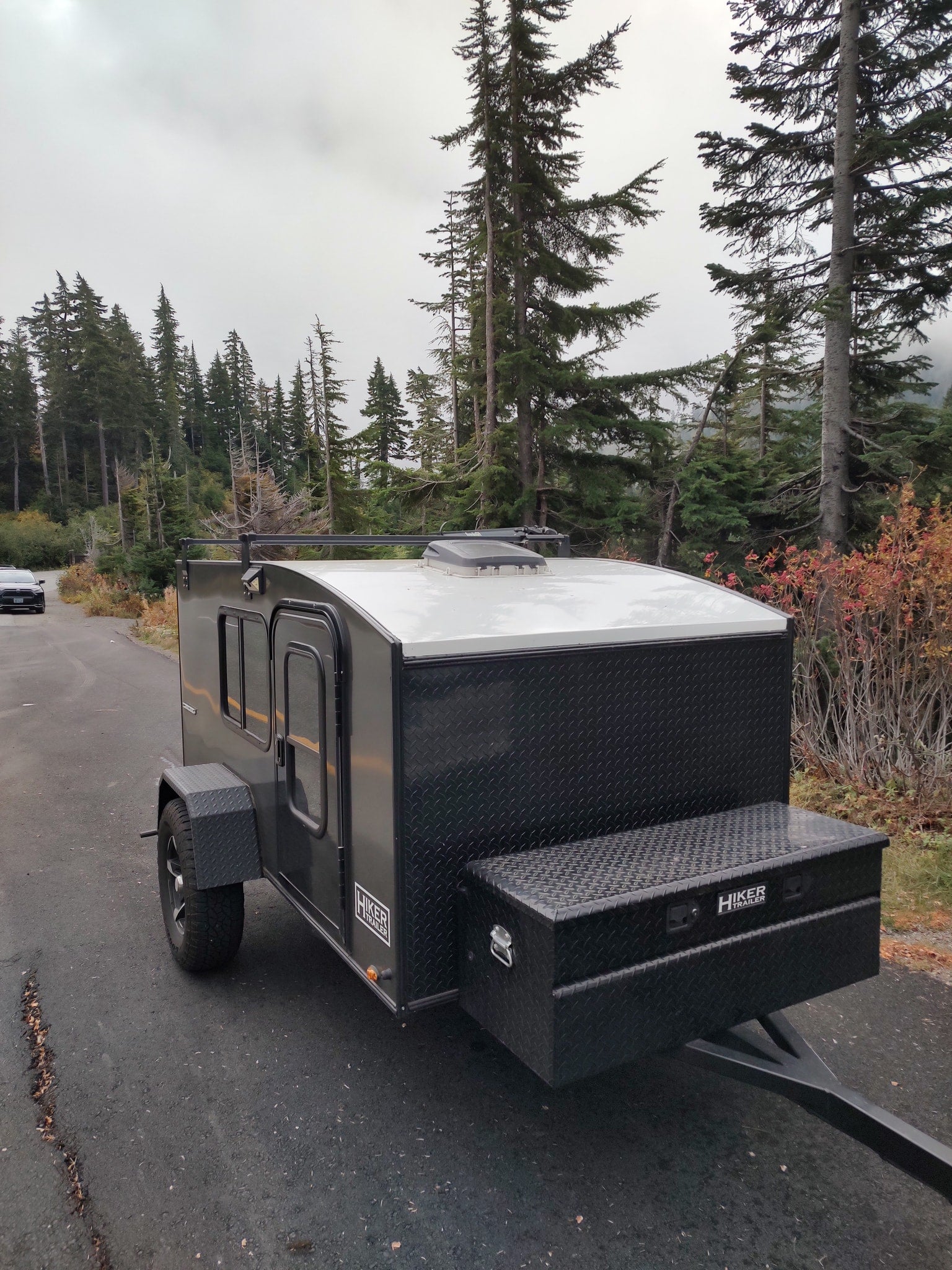 Compact Hiker camping trailer RV rental on a road through a dense evergreen forest. Perfect for outdoor travel.