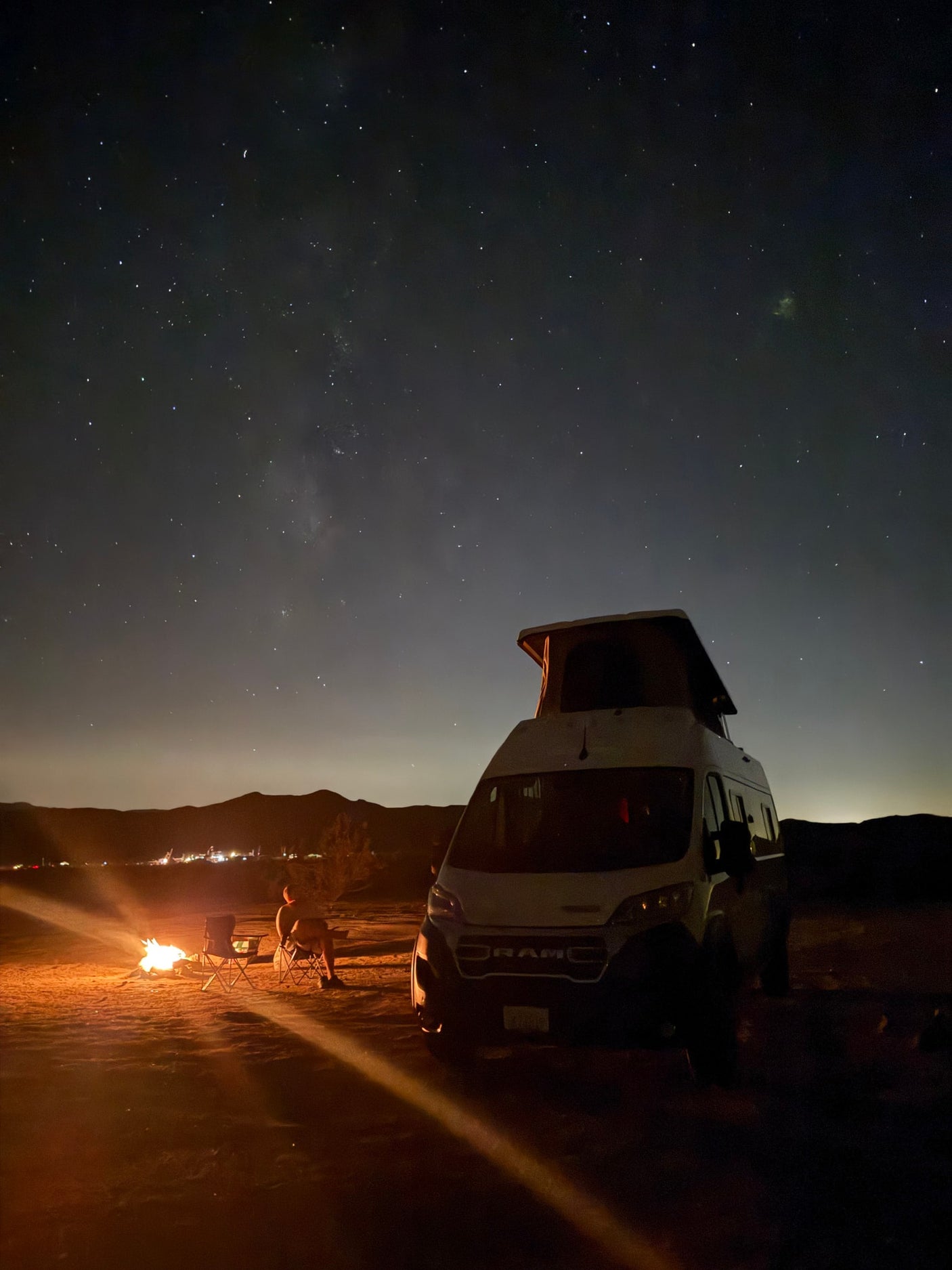 parked in a vast desert landscape at sunset.|A white RV rental camper van with a pop-top roof is set up for desert camping at sunset. Chairs await.|White Winnebago Solis RV rental van parked in a quiet desert setting with mountains at sunset.|Serene desert camping with a white RV rental. Pop-top extended