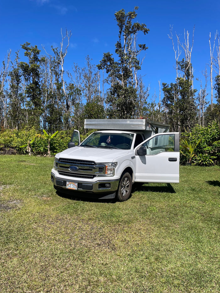 between a blue house and lush plants.|White Ford truck with rv rental camper on Hawaii's rocky coast