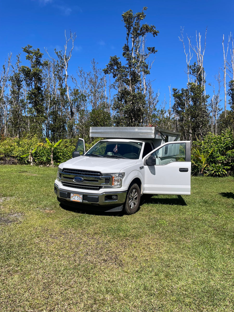 between a blue house and lush plants.|White Ford truck with rv rental camper on Hawaii's rocky coast