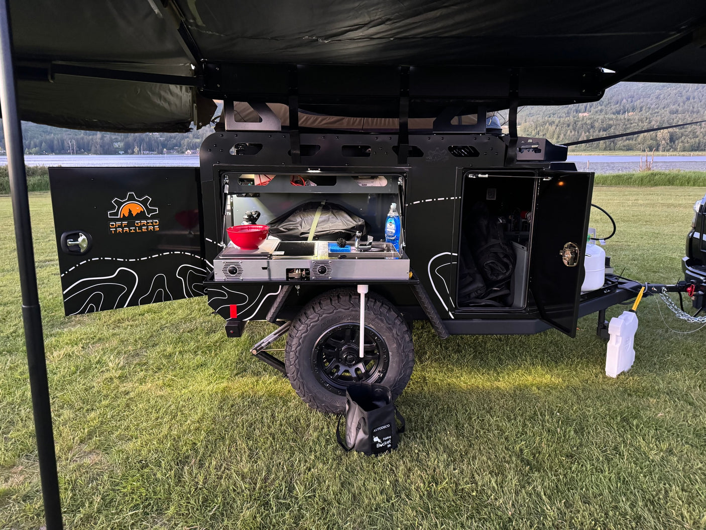 This off-grid trailer for rv rental features an open side revealing a well-equipped outdoor kitchen, parked on grass beside a tranquil lake.