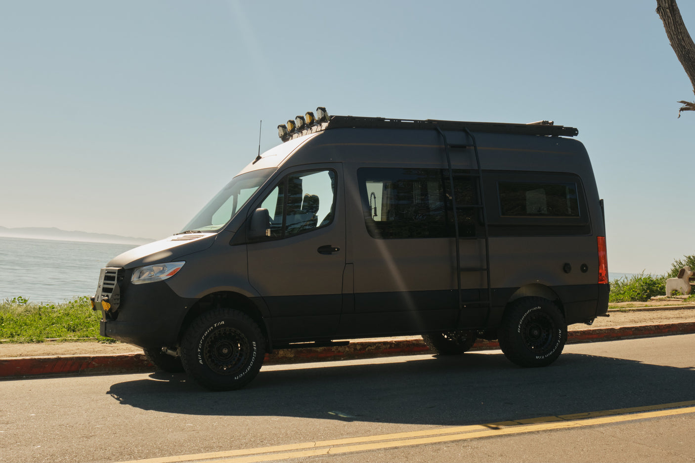 Off-road camper van RV rental, dark gray, with roof lights and ladder, parked by the ocean on a sunny day.