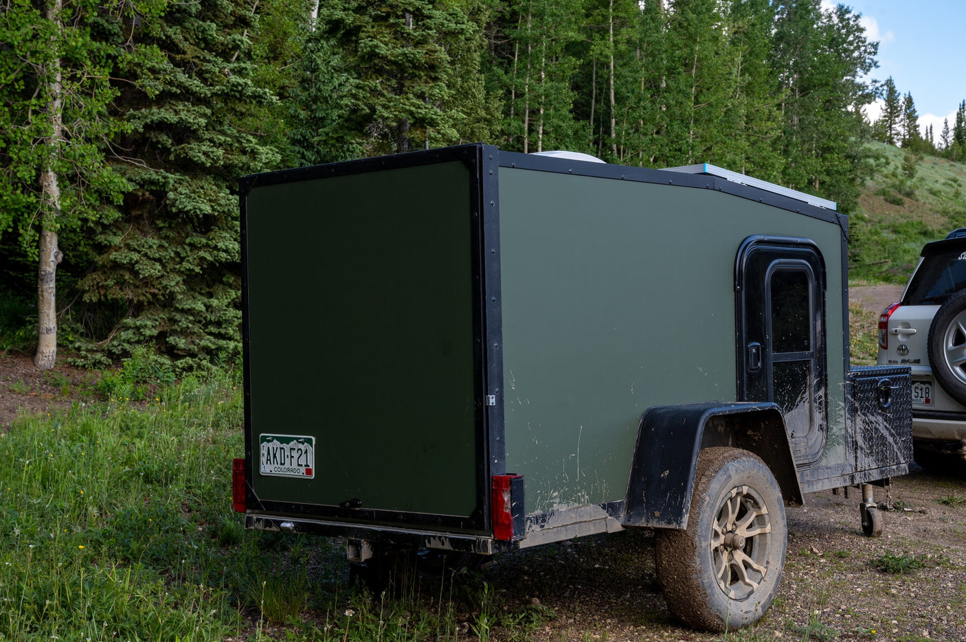 Mud-splattered green off-road camper trailer for RV rental, parked in a lush forest clearing. Ready for rugged adventures.