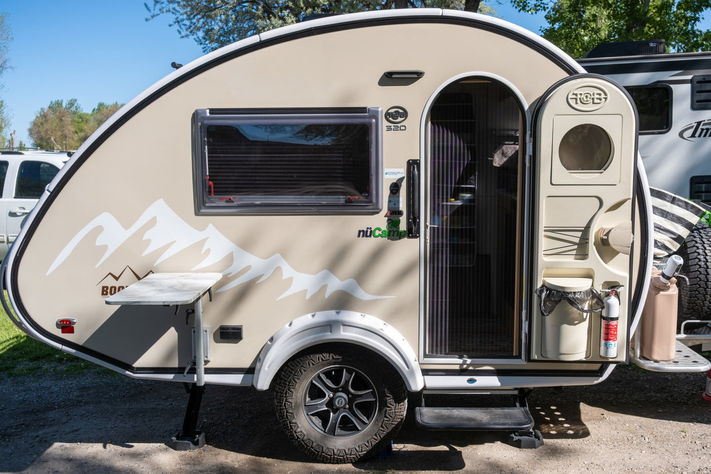 This beige nuCamp T@B 320 teardrop camper is ideal for an RV rental. Features a side table, open door, & mountain graphics.