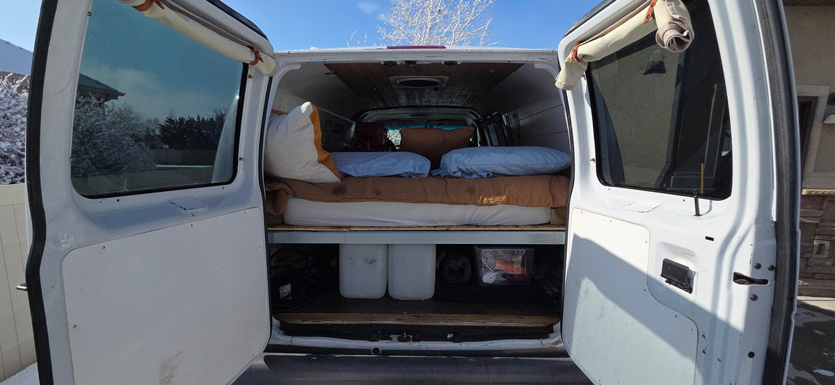 RV rental van interior with a bed, under-bed storage, and open rear doors revealing a snowy landscape outside. — photo 8