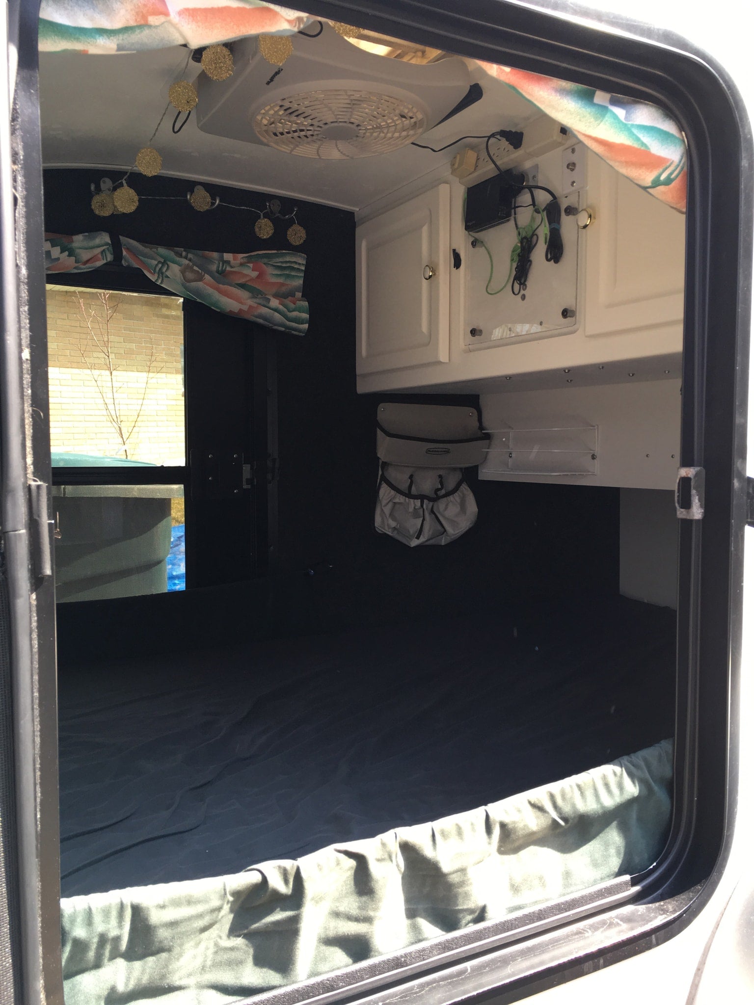 Inside a cozy RV rental: bed, overhead storage, fan, and string lights, ready for adventure.