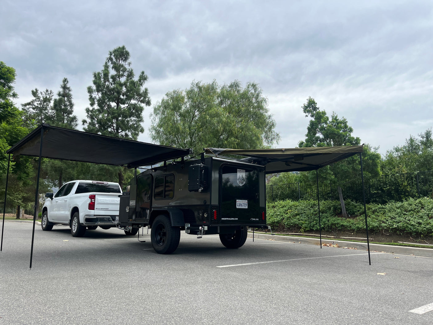 RV rental: White pickup truck and dark green camper trailer with extended awnings set up in a parking lot under a cloudy sky.