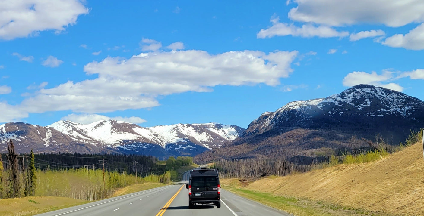 RV rental van driving on a scenic highway towards majestic snow-capped mountains under a clear blue sky.