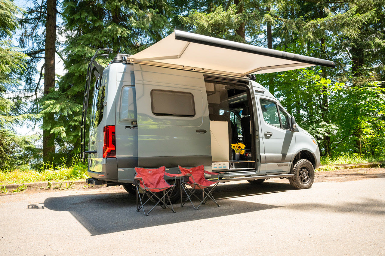Grey Winnebago RV rental parked in a sunny forest. An awning extends, side door is open with sunflowers, and two chairs are out.