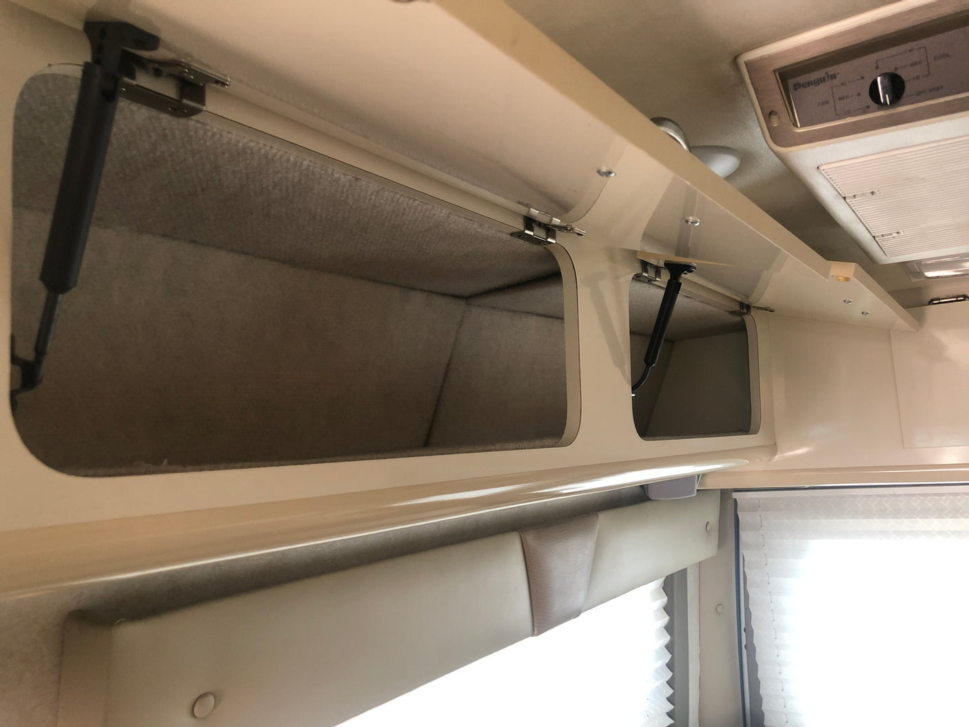Overhead cabinets with soft-lined interiors and gas struts in an RV rental. An AC unit and window with pleated blinds are also seen.