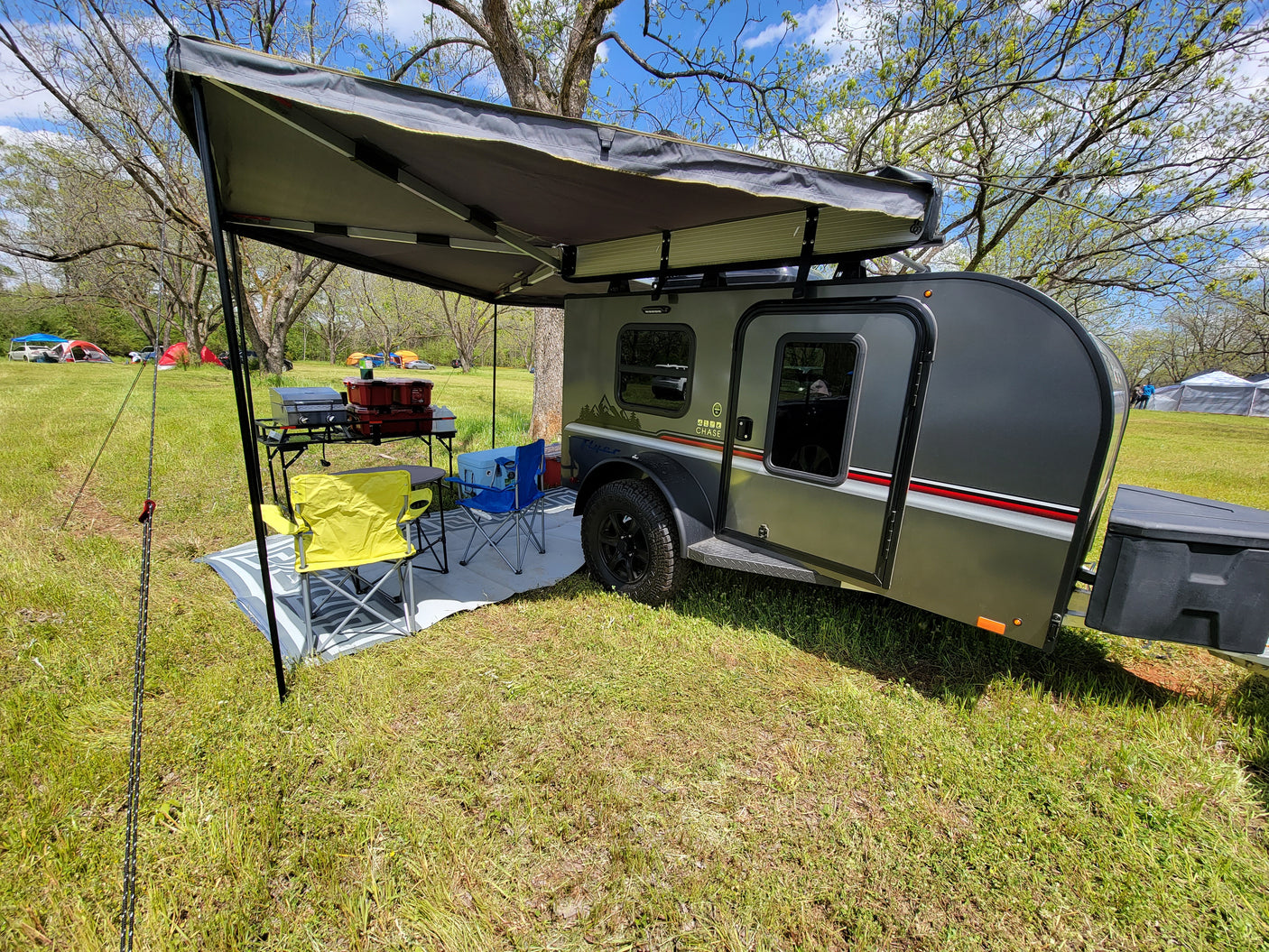 Explore nature with this compact RV rental! Teardrop trailer with awning, chairs & kitchen setup in a sunny campground.