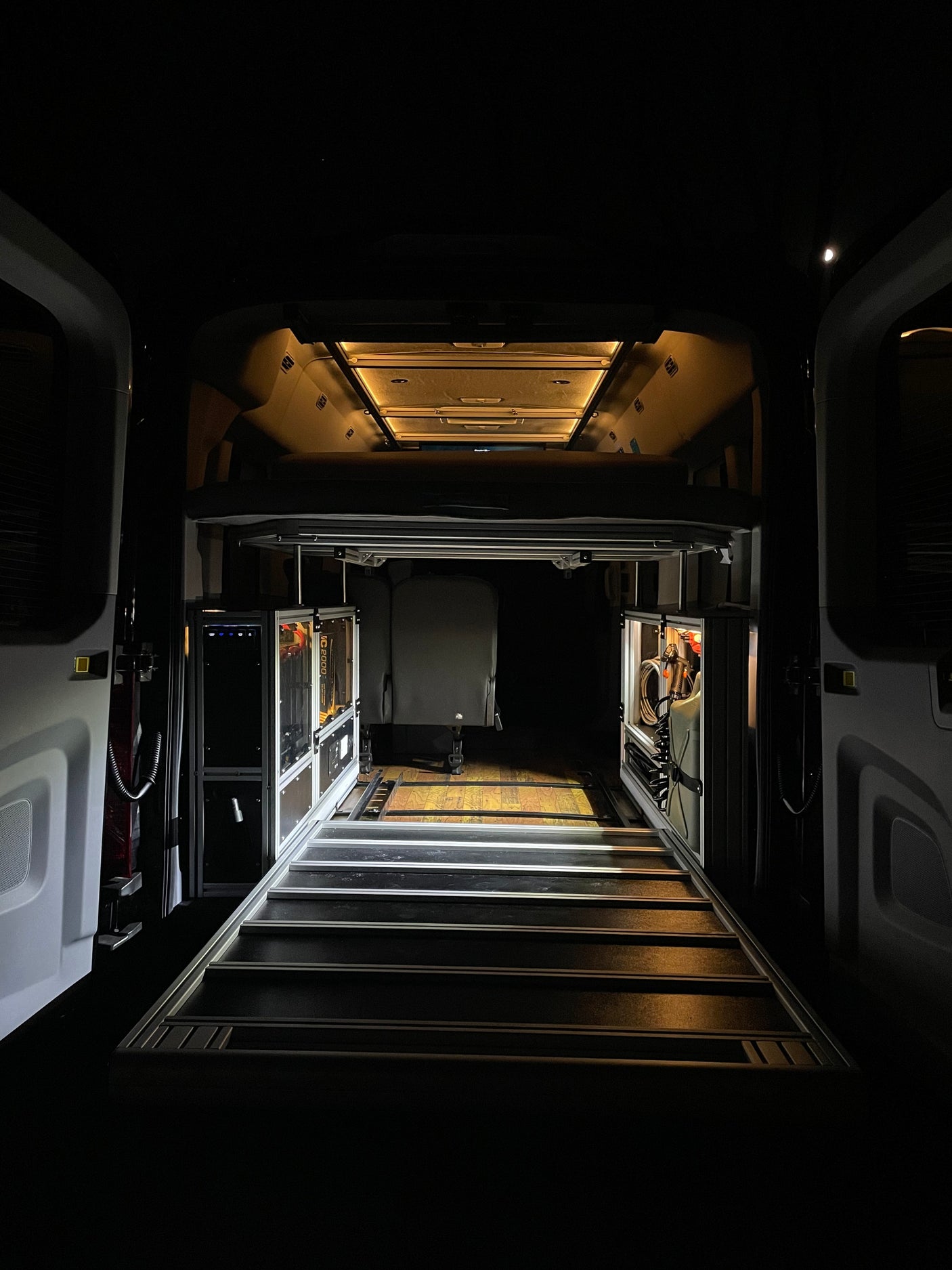Night view inside a custom rv rental camper van, revealing a raised bed, modular side storage with gear, and illuminated track flooring.