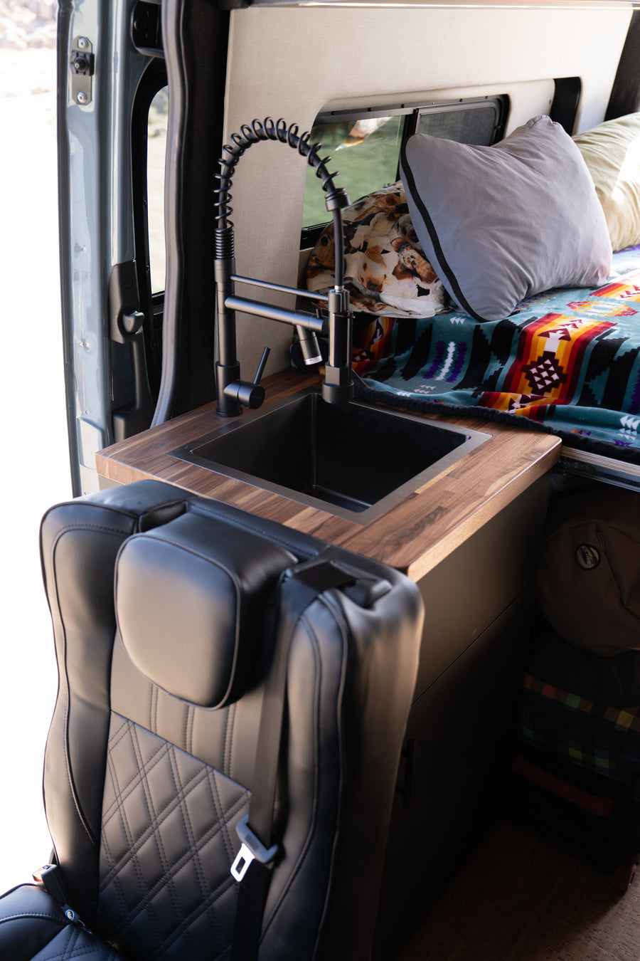 Experience an RV rental! Modern van interior featuring a sleek black sink, wooden counter, and quilted leather seat near the cozy bed.