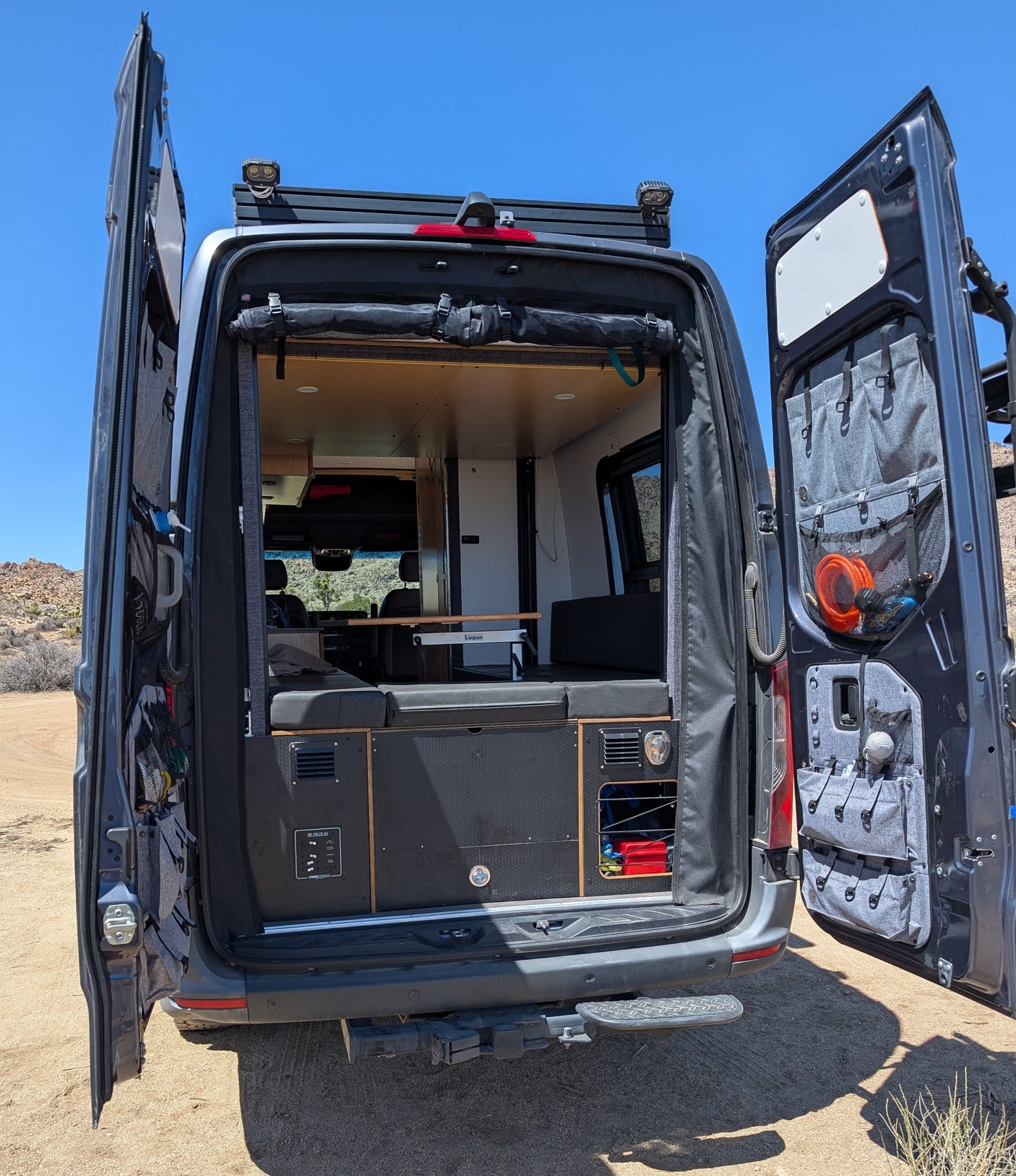 Rear view of an rv rental camper van with open doors, showcasing its custom interior, bed, table, and clever storage.