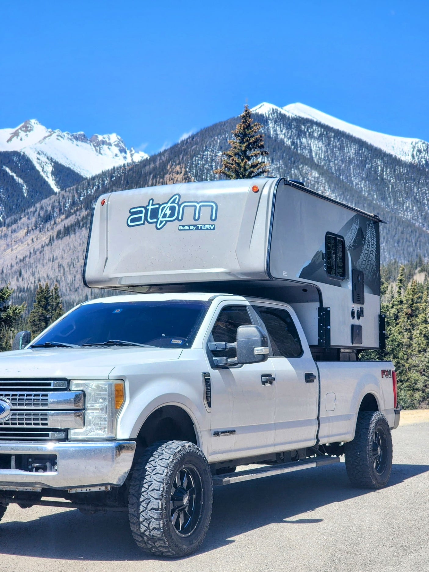 Experience mountain adventures with this RV rental: a white Ford pickup truck carrying an atøm truck camper against snowy peaks.