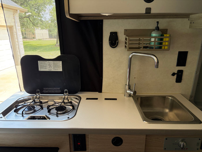RV rental kitchen: a two-burner stove, sink, faucet, and soap caddy on a light counter, with tank level controls.