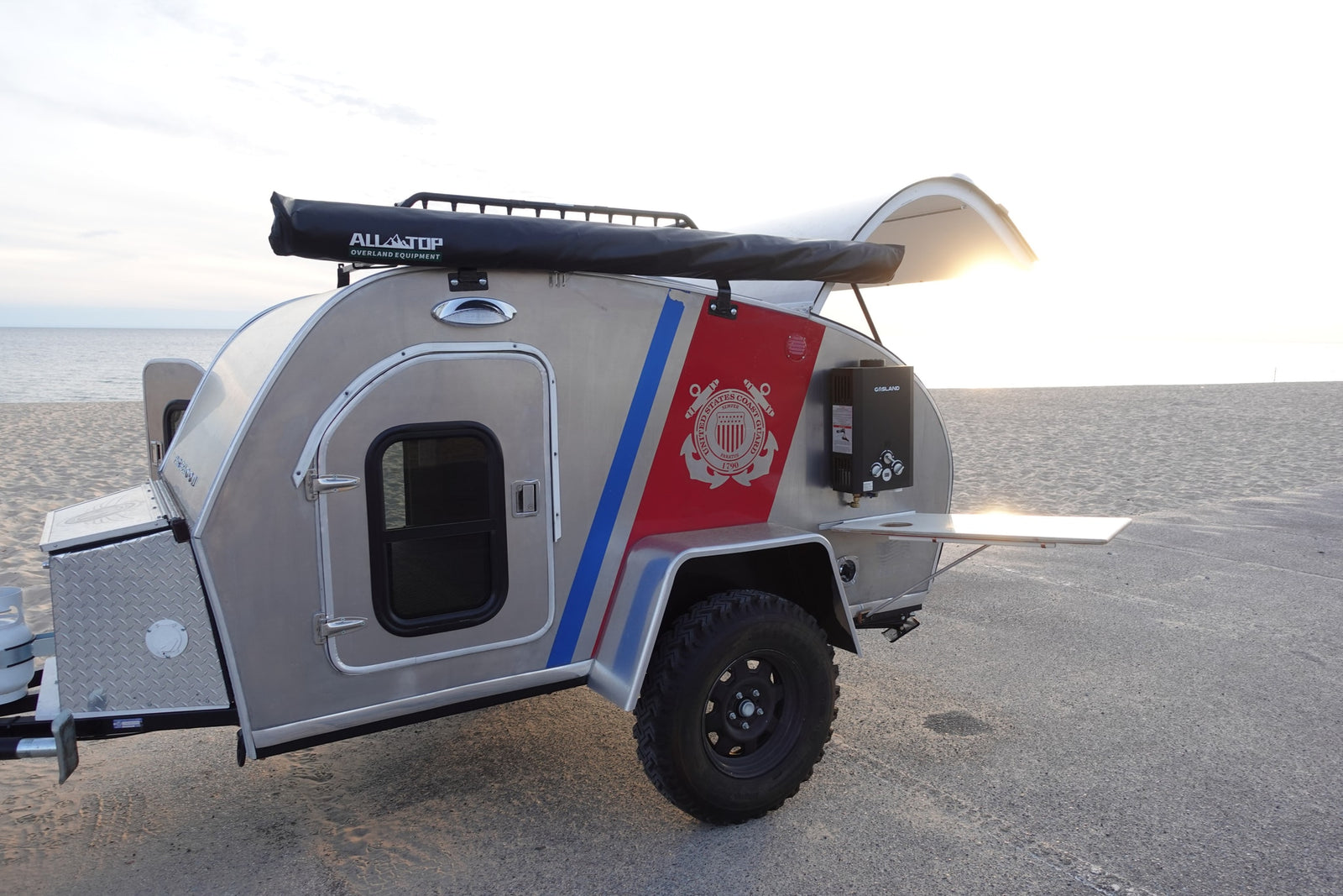 This unique teardrop RV rental, complete with an awning and Coast Guard theme, is parked on the beach for your next adventure.