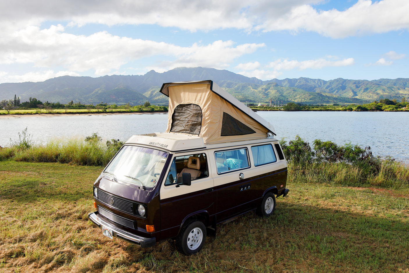 Brown & beige VW Westfalia pop-top camper van (RV rental) by a lake with scenic mountains. Ready for adventure!