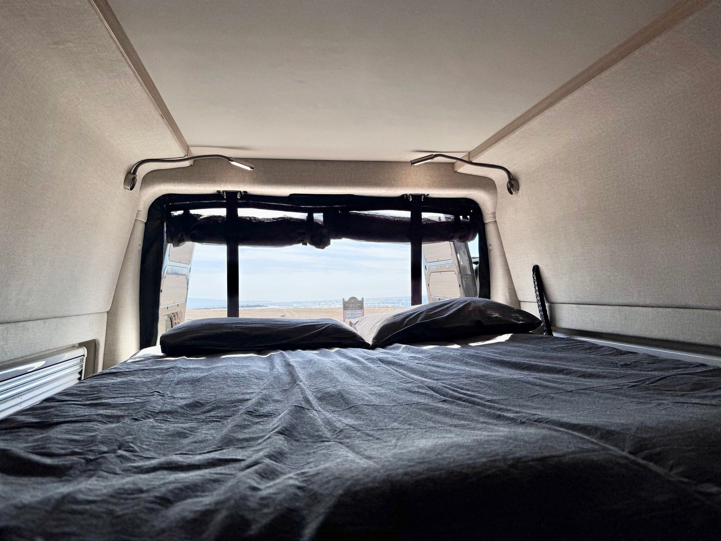 View from an **RV rental** bed, showing a cozy interior, pillows, and a sandy beach and ocean through the rear window.