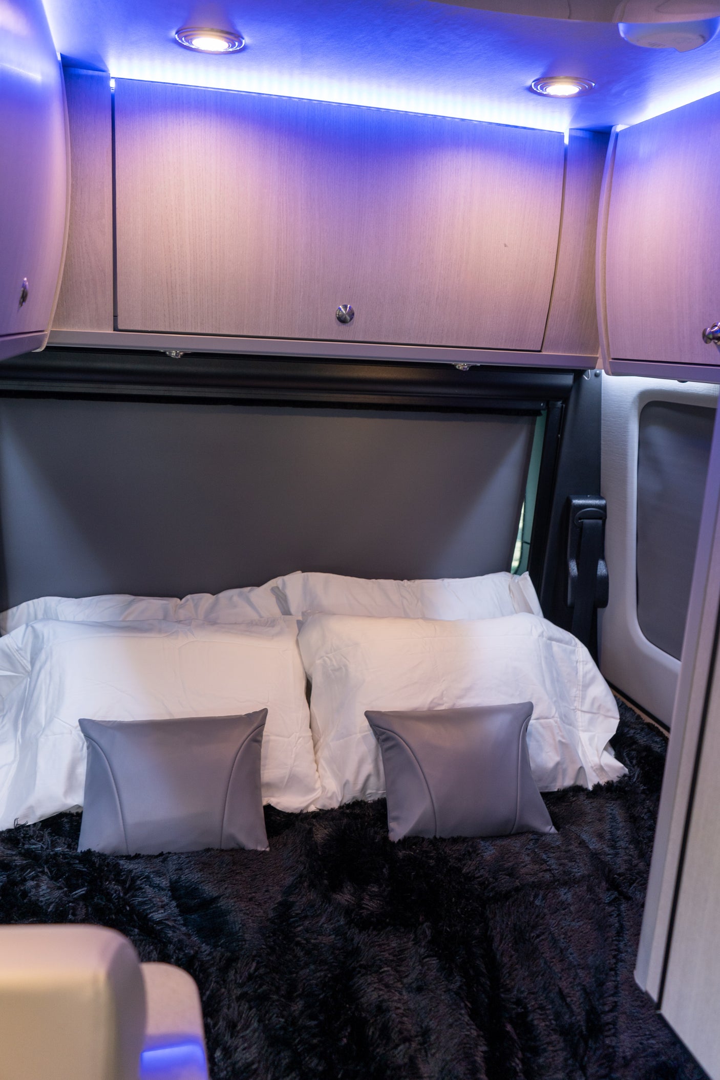 The bed area of an RV rental, featuring blue ambient lighting, overhead cabinets, white pillows, and a fluffy black blanket.