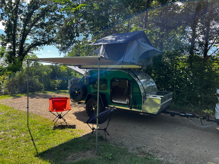 RV rental teardrop camper with a roof-top tent and extended awning, set up in a lush outdoor camping spot.