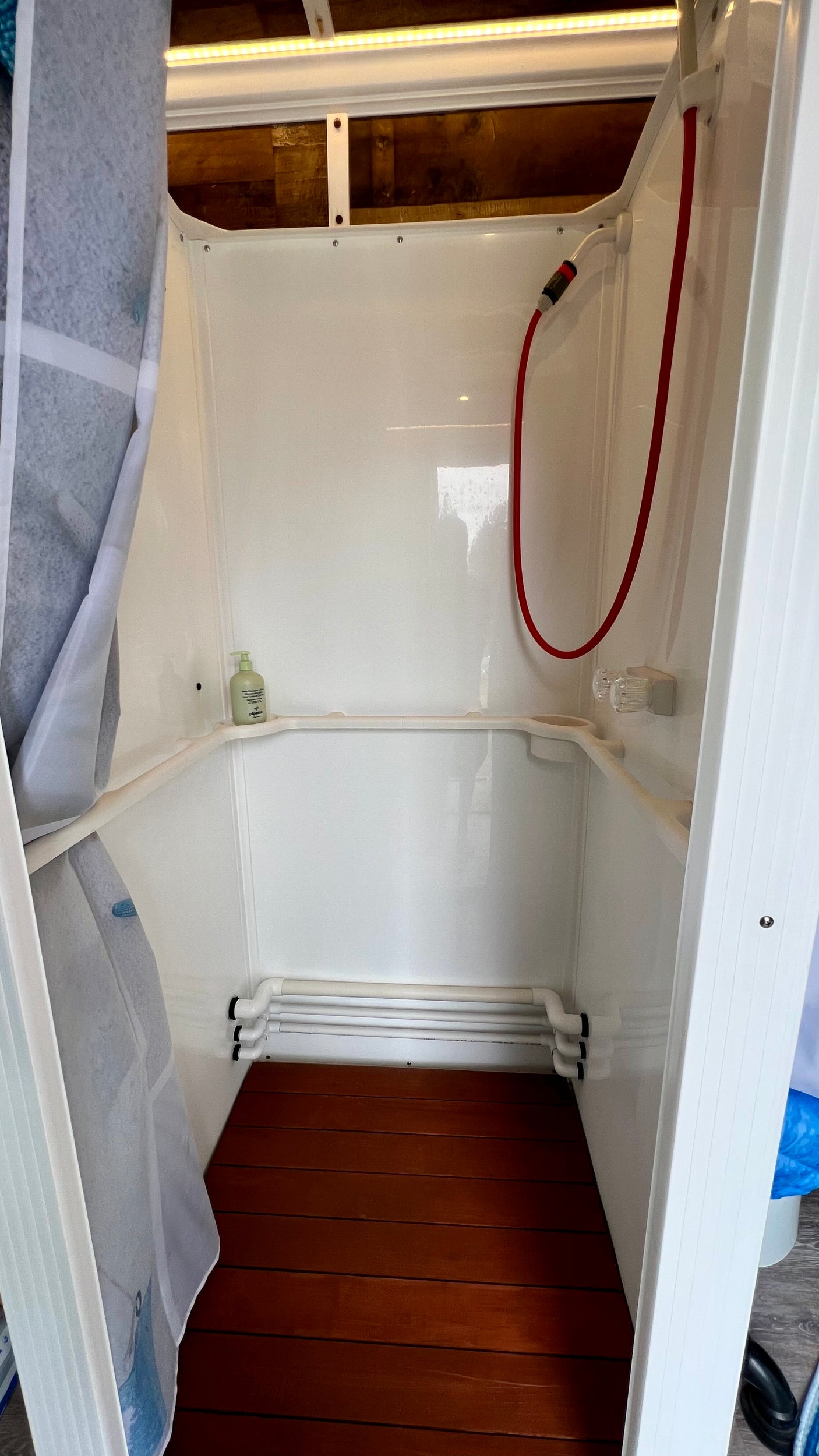 Compact white shower enclosure with red hose shower, wood floor, soap, in an rv rental.