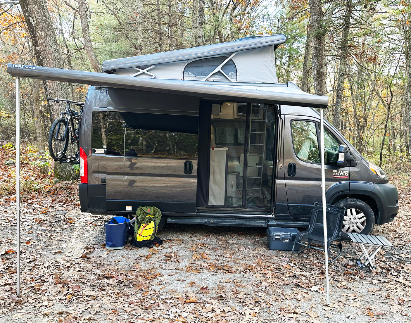 A grey pop-top RV rental with an extended Thule awning is set up in a leafy forest. A bike, chair, and table are visible.