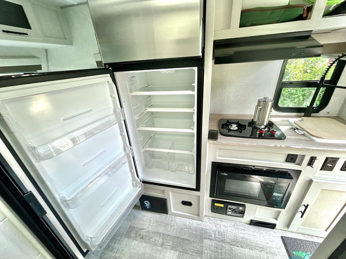 Inside an RV rental, an open fridge/freezer with clear shelves. The compact kitchen features a stove, microwave, and window.