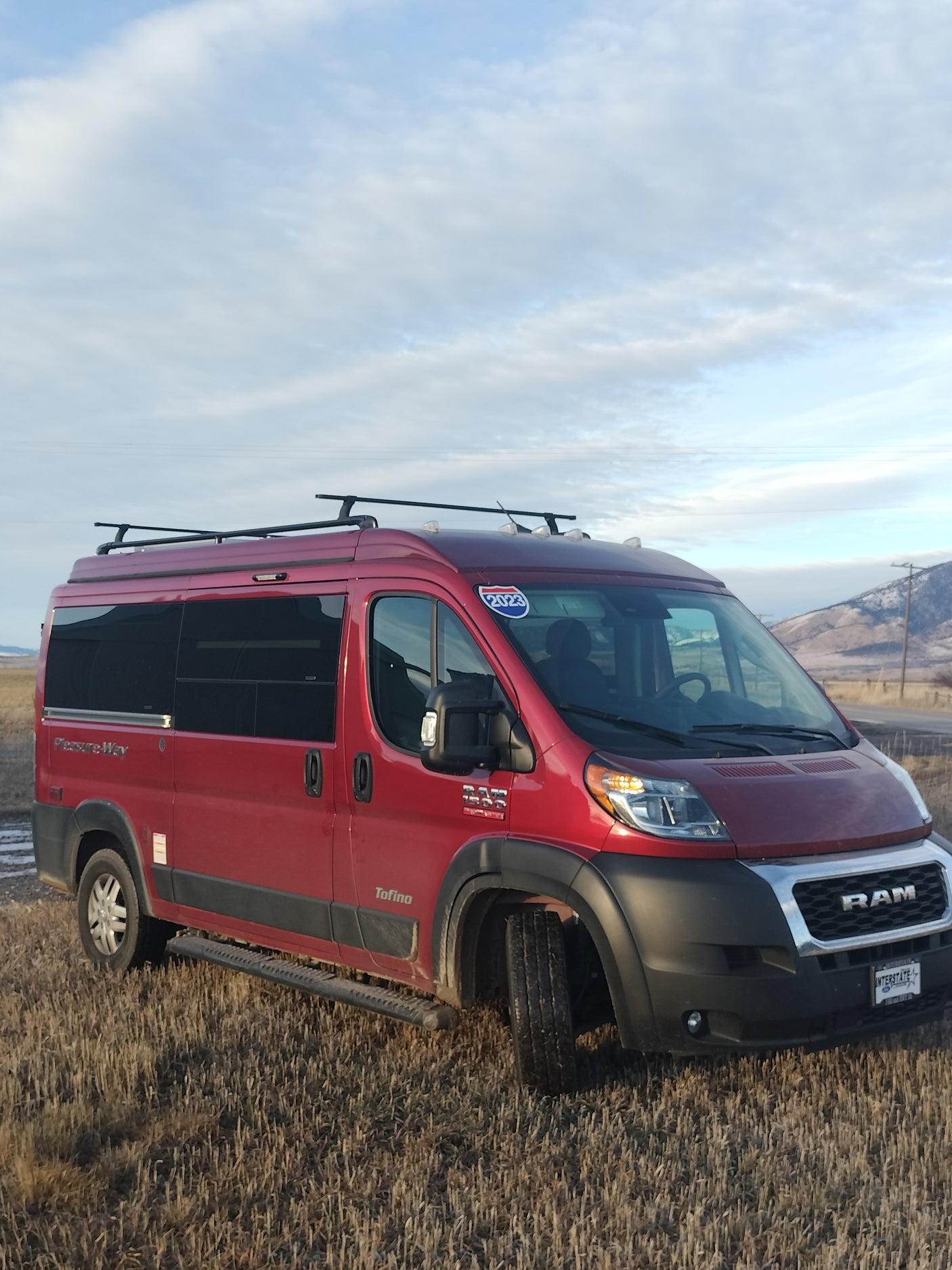 Explore landscapes with this red Pleasure-Way Tofino camper van, an ideal rv rental, parked in a dry field with mountains.