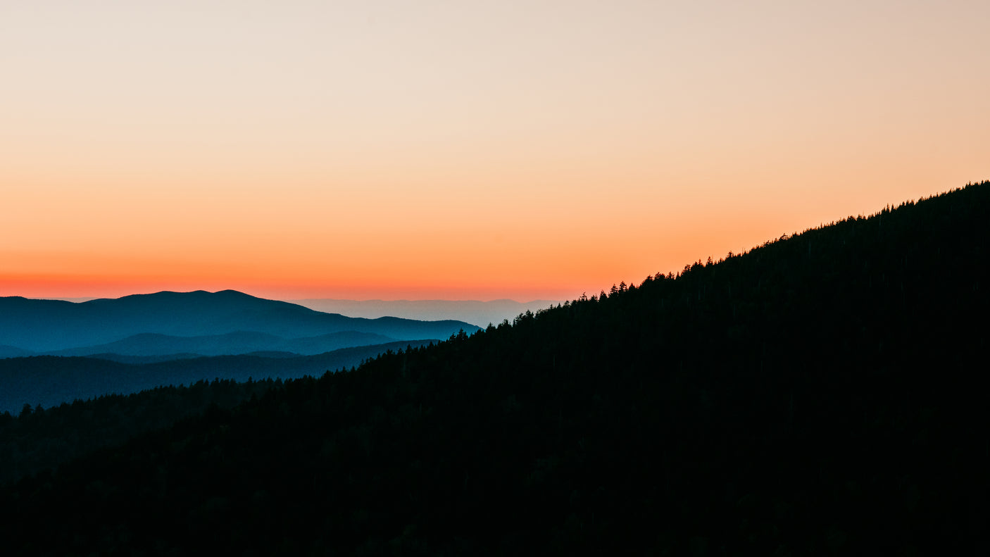Vibrant orange sunset illuminates distant blue mountain layers & dark foreground trees. Imagine this view from an RV rental.