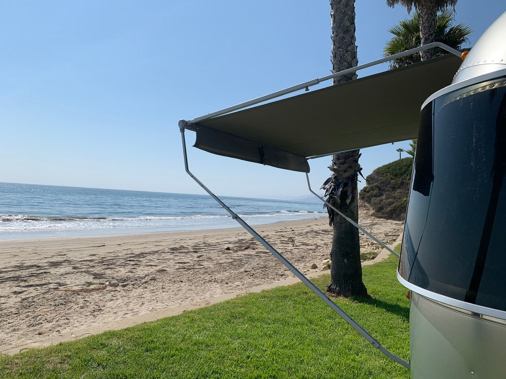 This RV rental offers a stunning beach view. An Airstream with awning extended, overlooking the ocean, sand, and palm trees.
