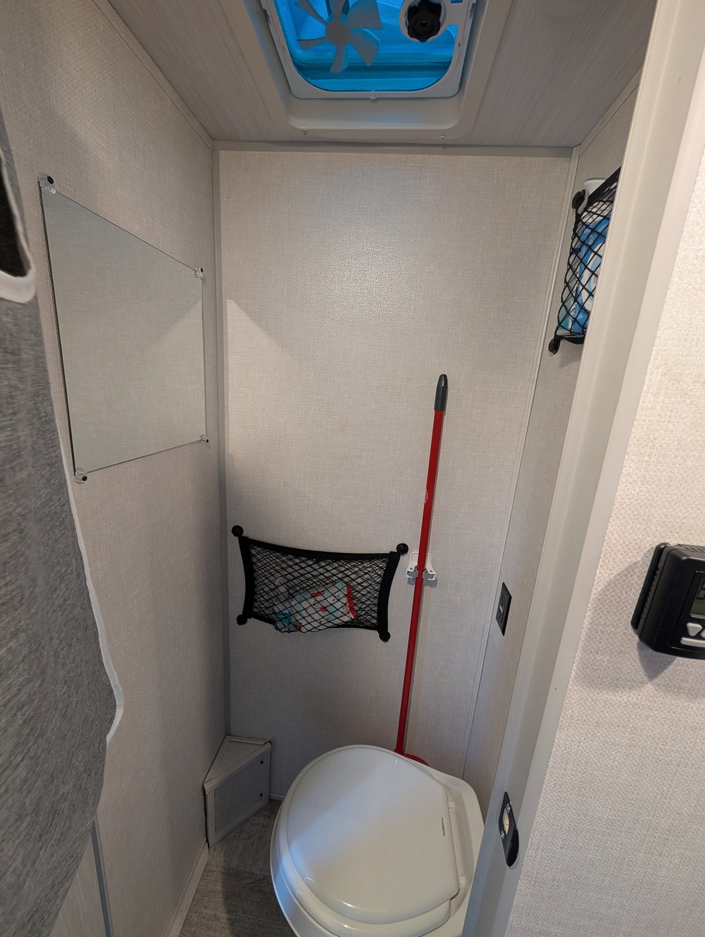 Inside an RV rental, this compact bathroom features a toilet, overhead fan, mirror, and wall storage with a broom.