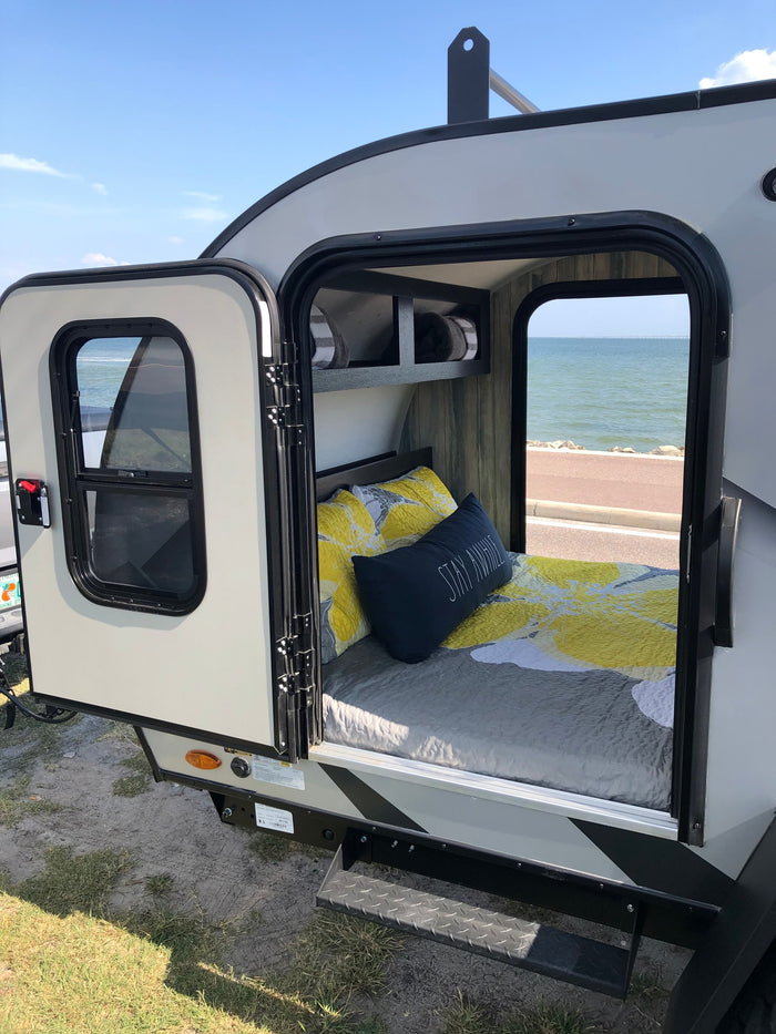 Cozy rv rental teardrop camper bedroom with 'Stay Awhile' pillow, open to ocean views and coastal road.