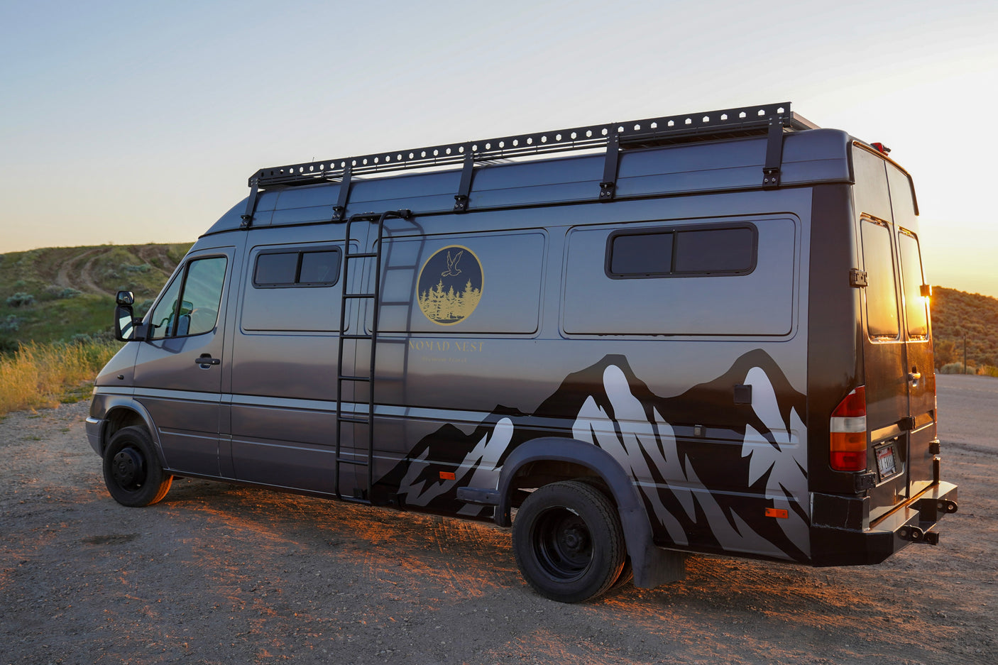 This custom gray Nomad Nest **rv rental** van, featuring mountain graphics and a roof rack, is parked on a dirt road at sunset.