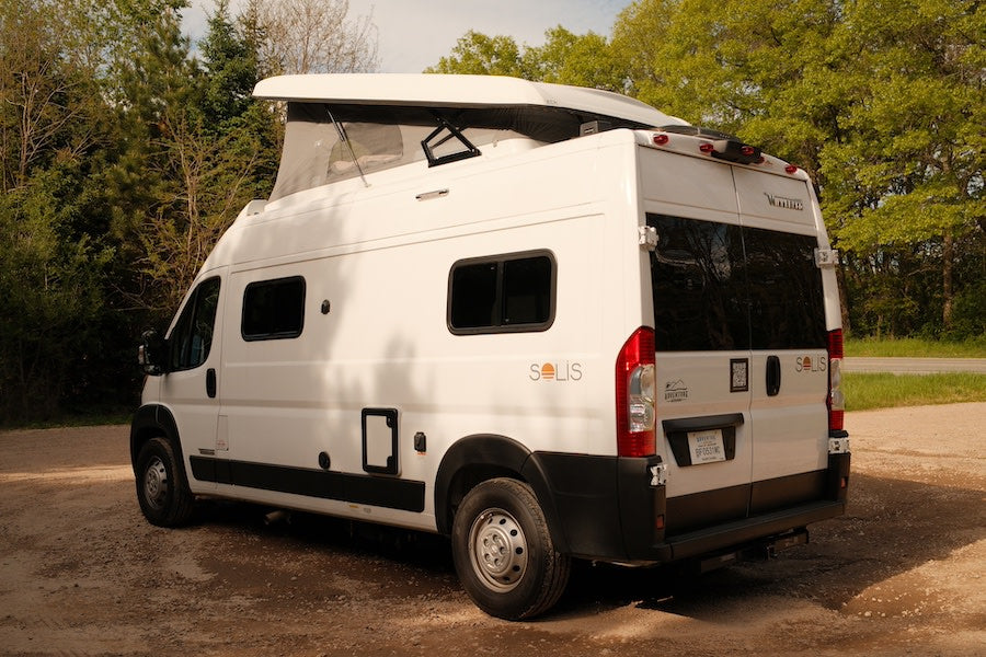 White Winnebago Solis pop-top camper van rental parked on a dirt path amidst trees, perfect for outdoor trips.